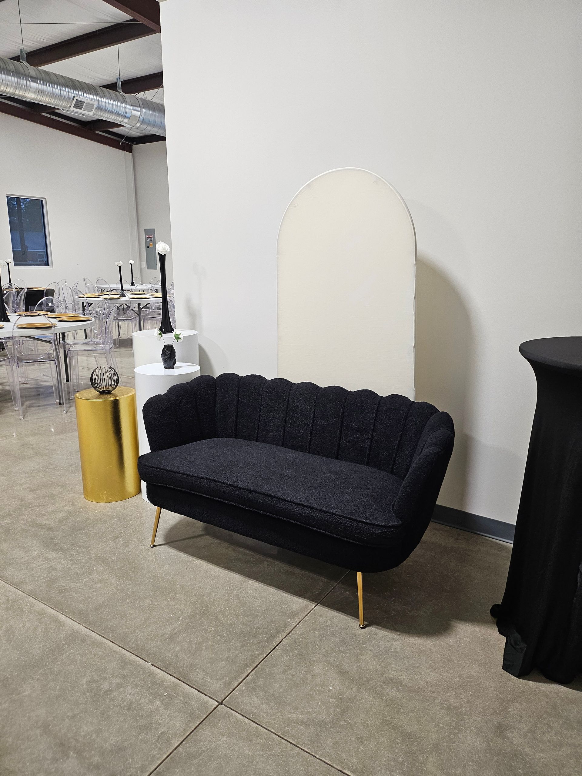 Black loveseat against a white wall in an industrial-style room with concrete floor and tables in the background