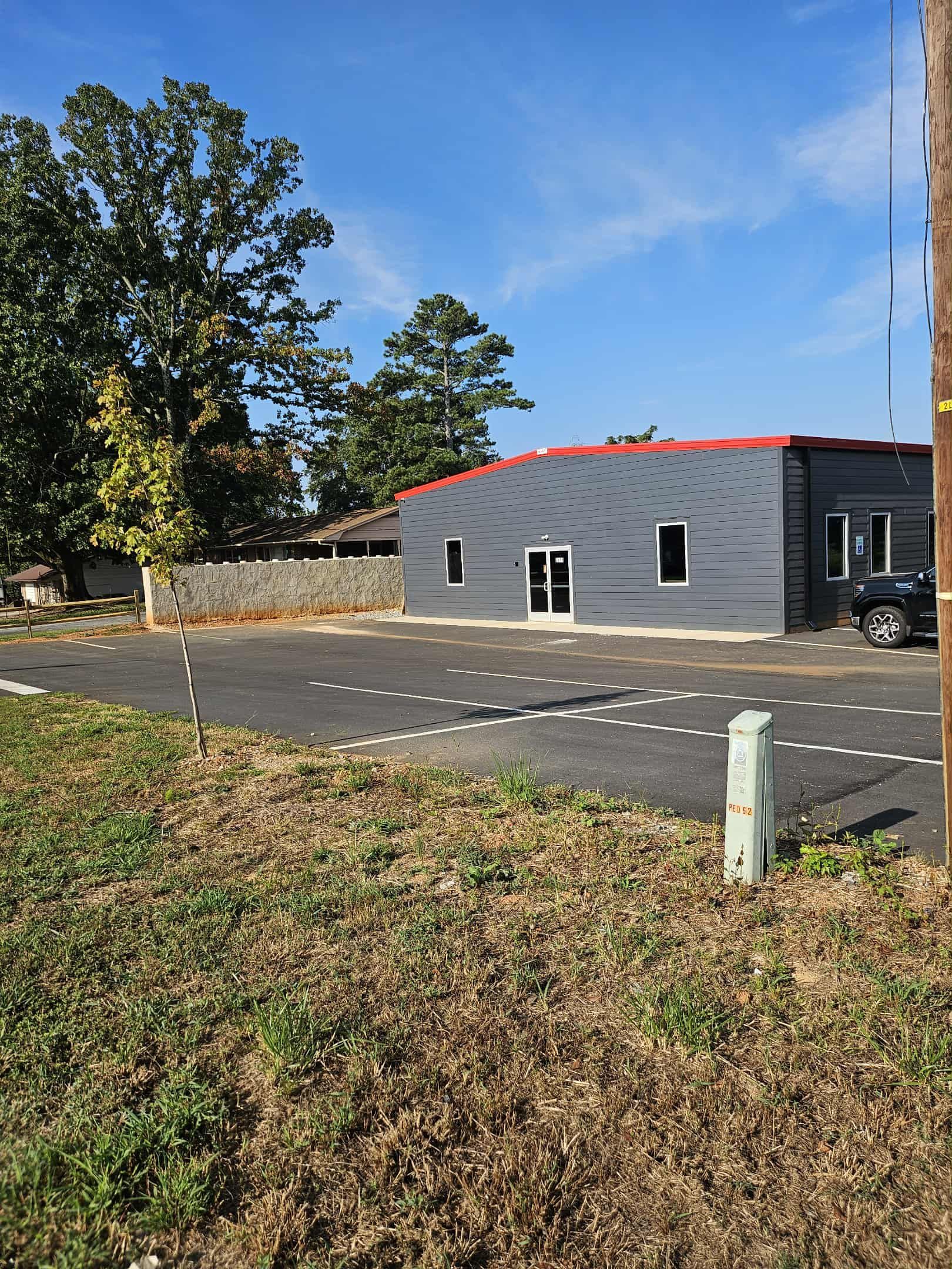 Small gray building by a road, with trees, grassy foreground, and blue sky.