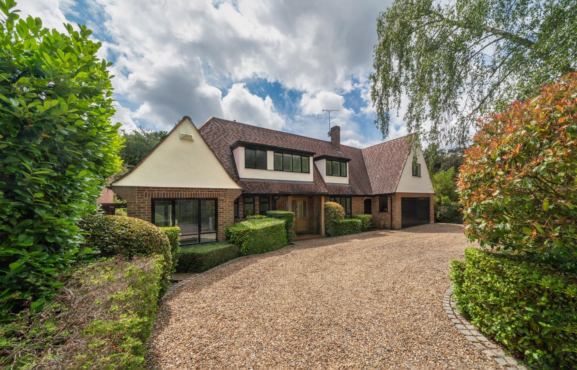 Large brick house with a gravel driveway, surrounded by lush green trees and a partly cloudy sky
