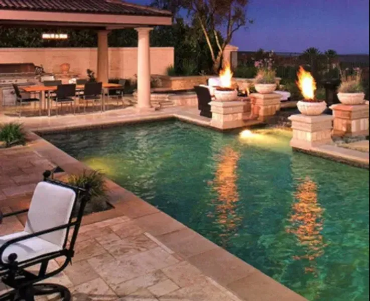 Outdoor pool at dusk with turquoise water, stone patio, lounge chairs, and a lit pergola seating area.