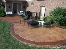 Curved stamped-concrete patio with chairs and table beside a brick house and garden bed