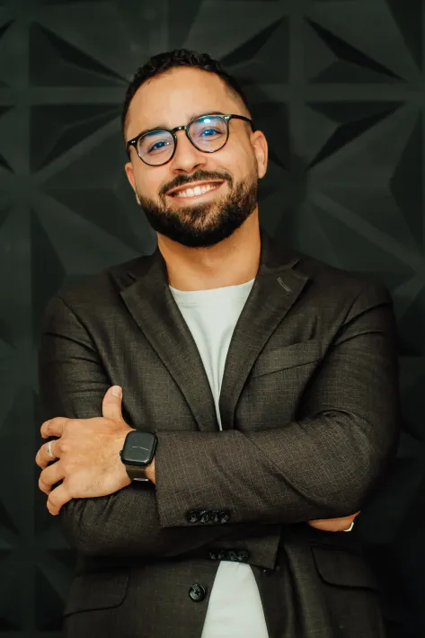 Man with glasses and beard, smiles with arms crossed, wearing a blazer, black background.