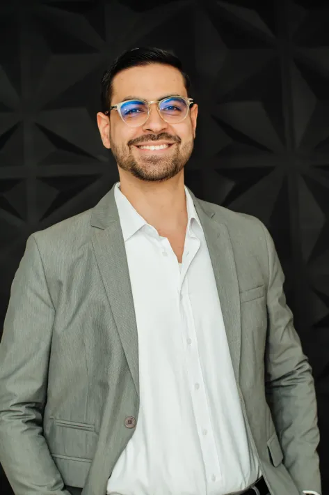 Man in a gray blazer, white shirt, and glasses smiling in front of a black textured wall.