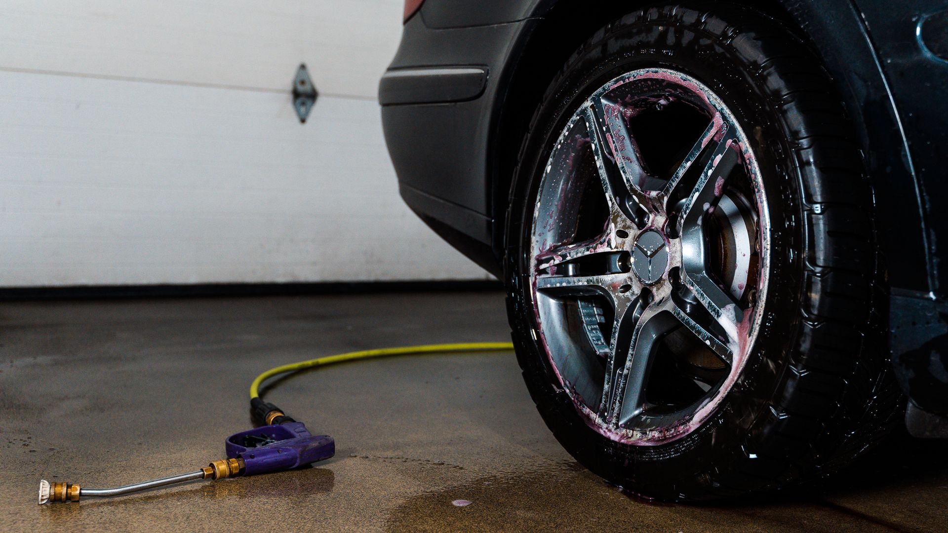 A spray nozzle attached to a yellow hose lies on a wet garage floor next to a soapy car tire and rim.