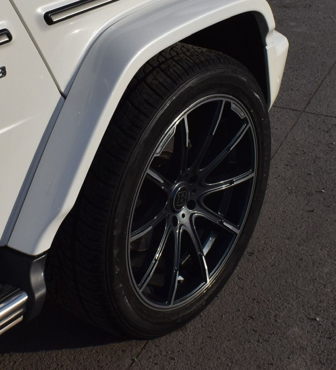 A close-up of a black multi-spoke wheel on a white Mercedes-Benz G-Class SUV parked on asphalt.