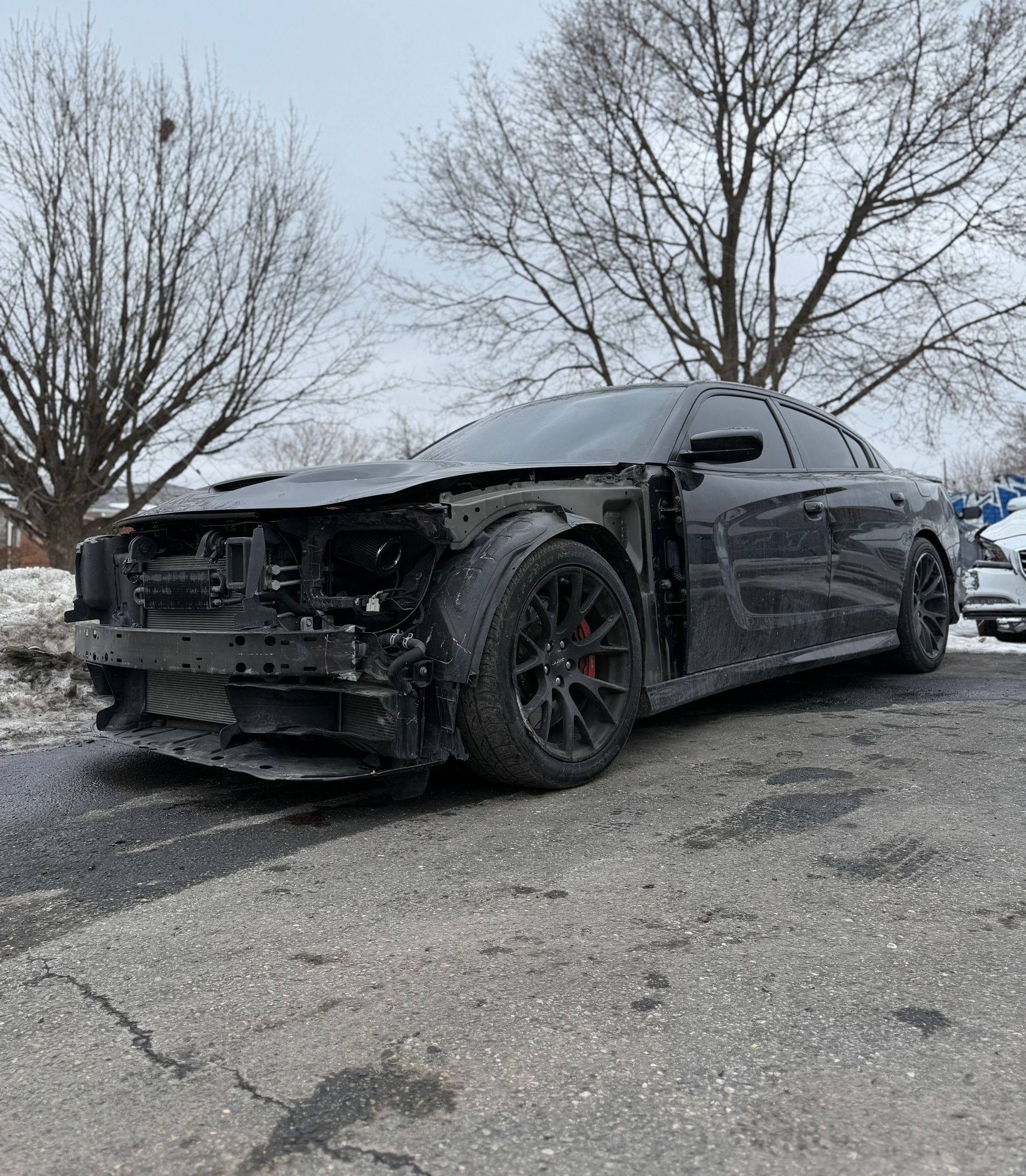 A damaged black Dodge Charger with its front bumper and headlight assembly missing, parked on an asphalt lot in winter.