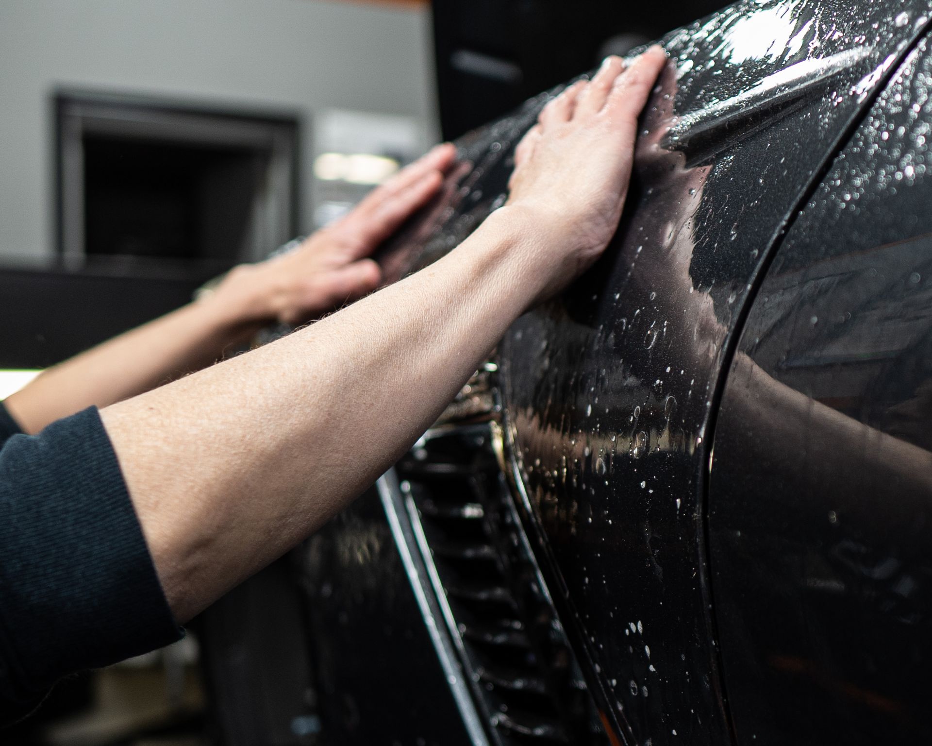 A person's hands smoothing a clear protective film onto the black paint of a car.