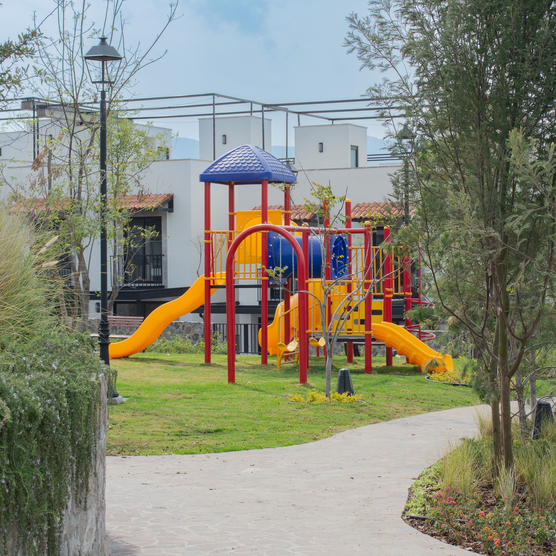 Una colorida estructura de juegos
infantiles con toboganes y techo azul, situada sobre un césped junto a un sendero y edificios blancos.