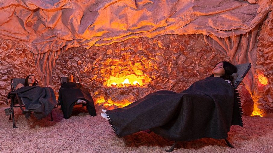 People relaxing in a salt cave; reddish-orange lighting, seated on recliners, draped in black blankets.