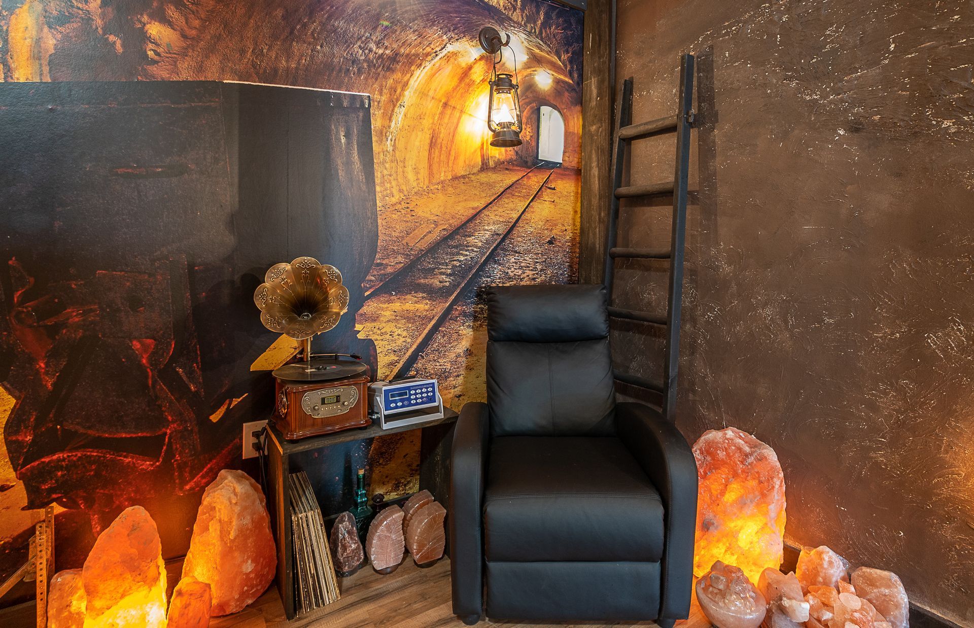 Cozy room with black recliner, themed decor. A train tunnel mural, vintage record player, and salt lamps glow.