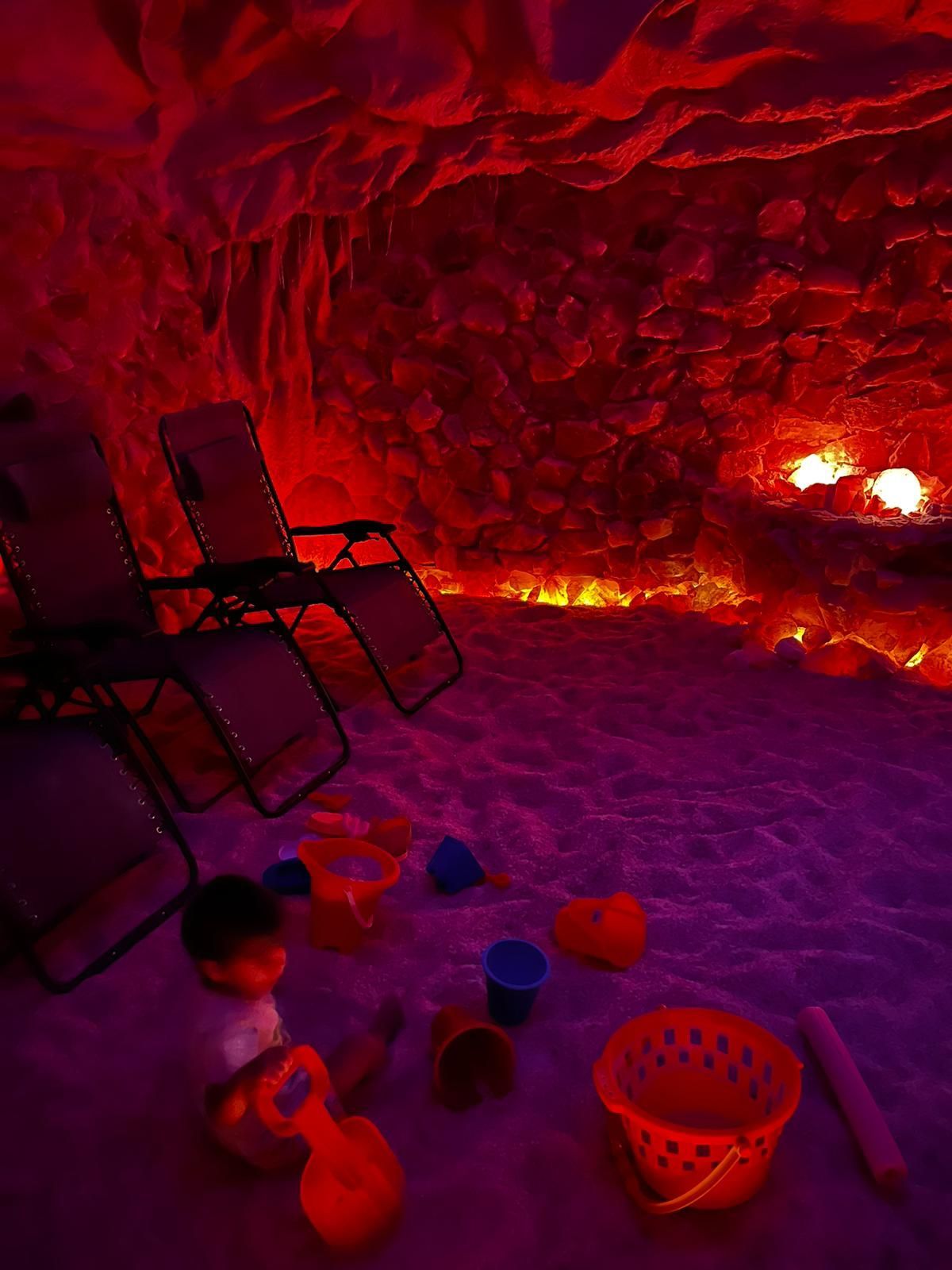 A dimly lit salt cave with red-orange walls, beach chairs, and children’s toys on the salt floor.