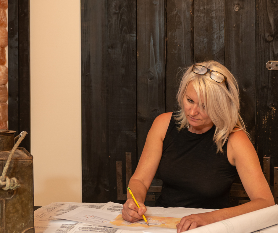Woman with blonde hair, eyeglasses, sketching at a table with a yellow pencil. Dark wood background.