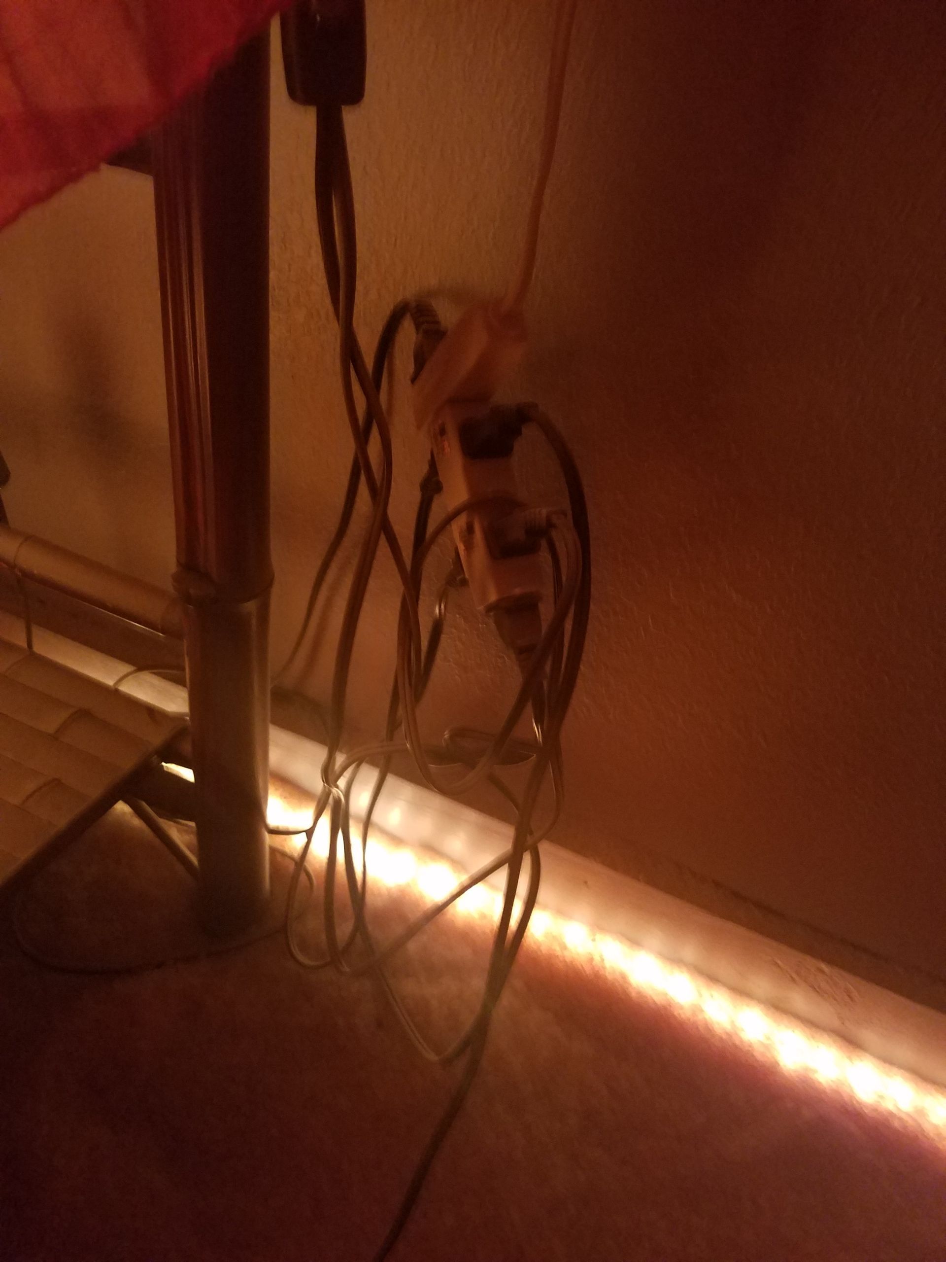 Messy tangle of electrical cords plugged into power outlets near a wooden floor with LED strip lighting.