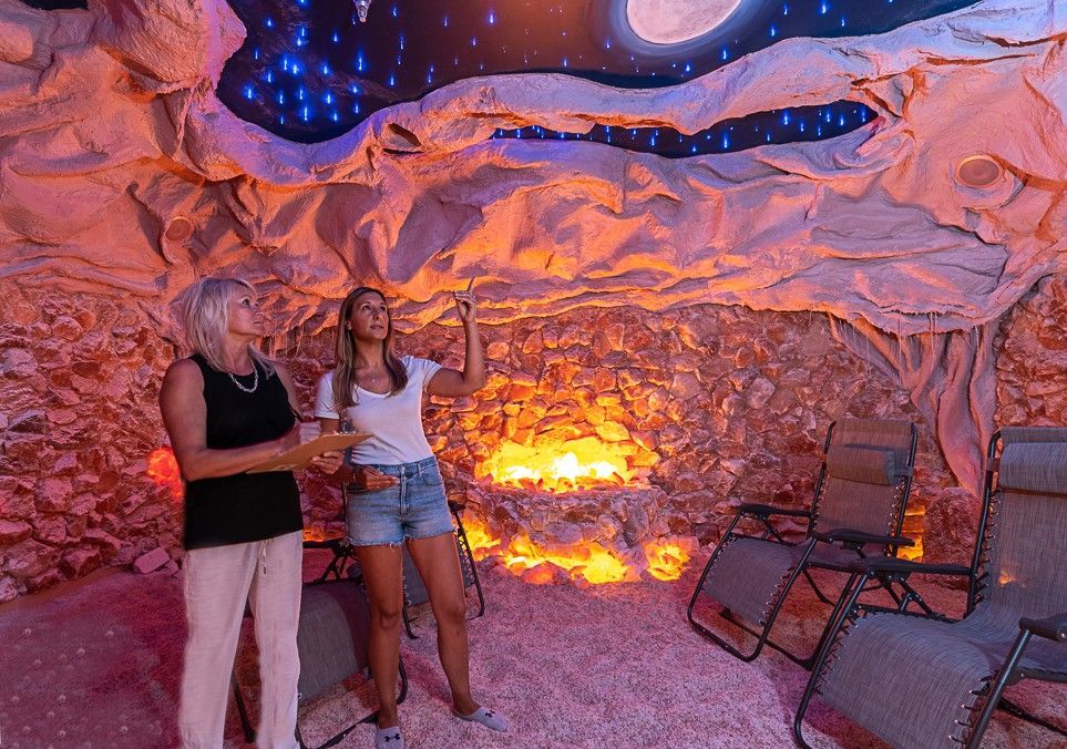 Two people in a salt cave, looking at lit ceiling. Pink salt walls, chairs, warm lighting.