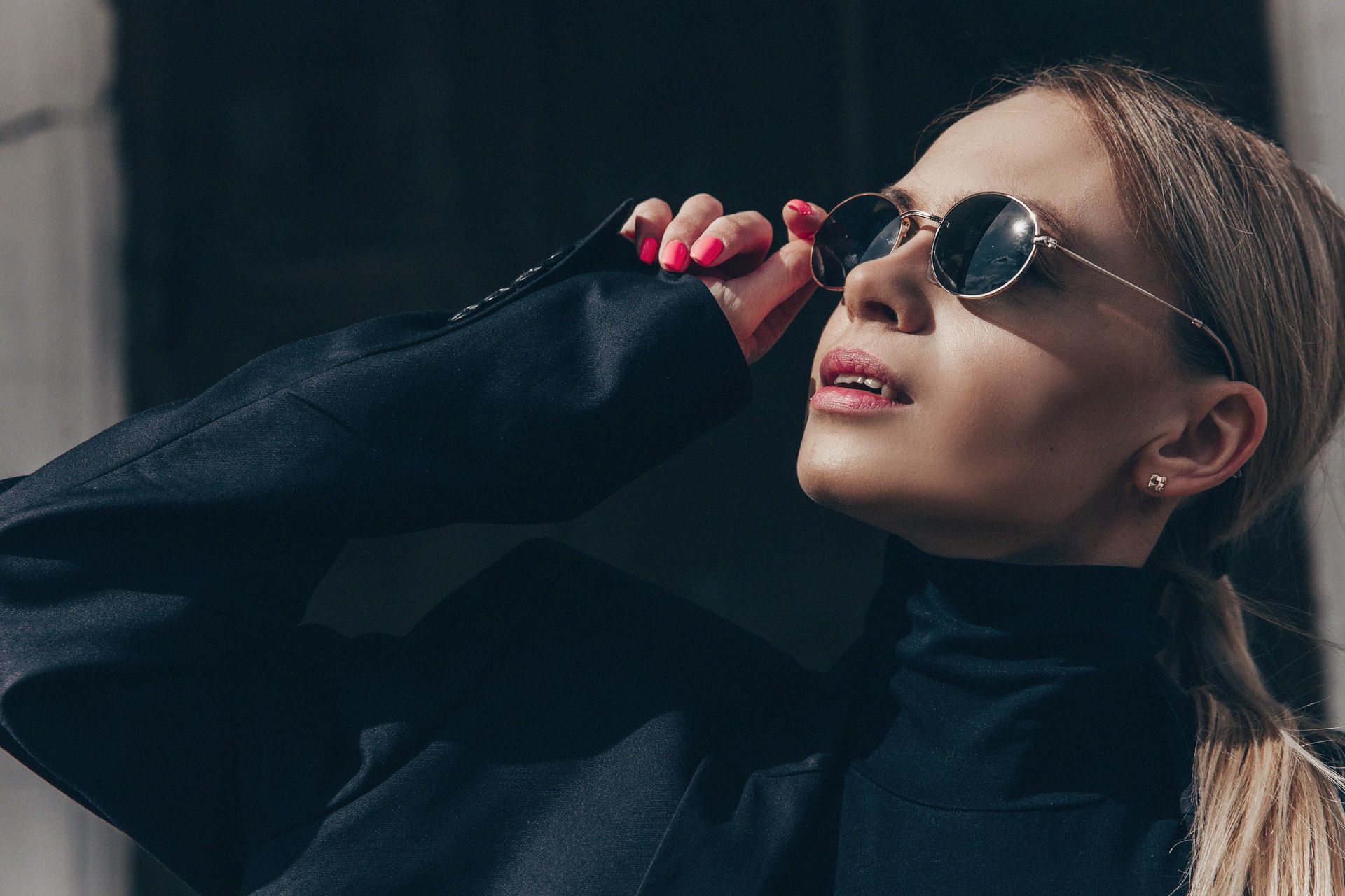 Une femme portant des lunettes de soleil et une veste noire se tient devant un bâtiment.