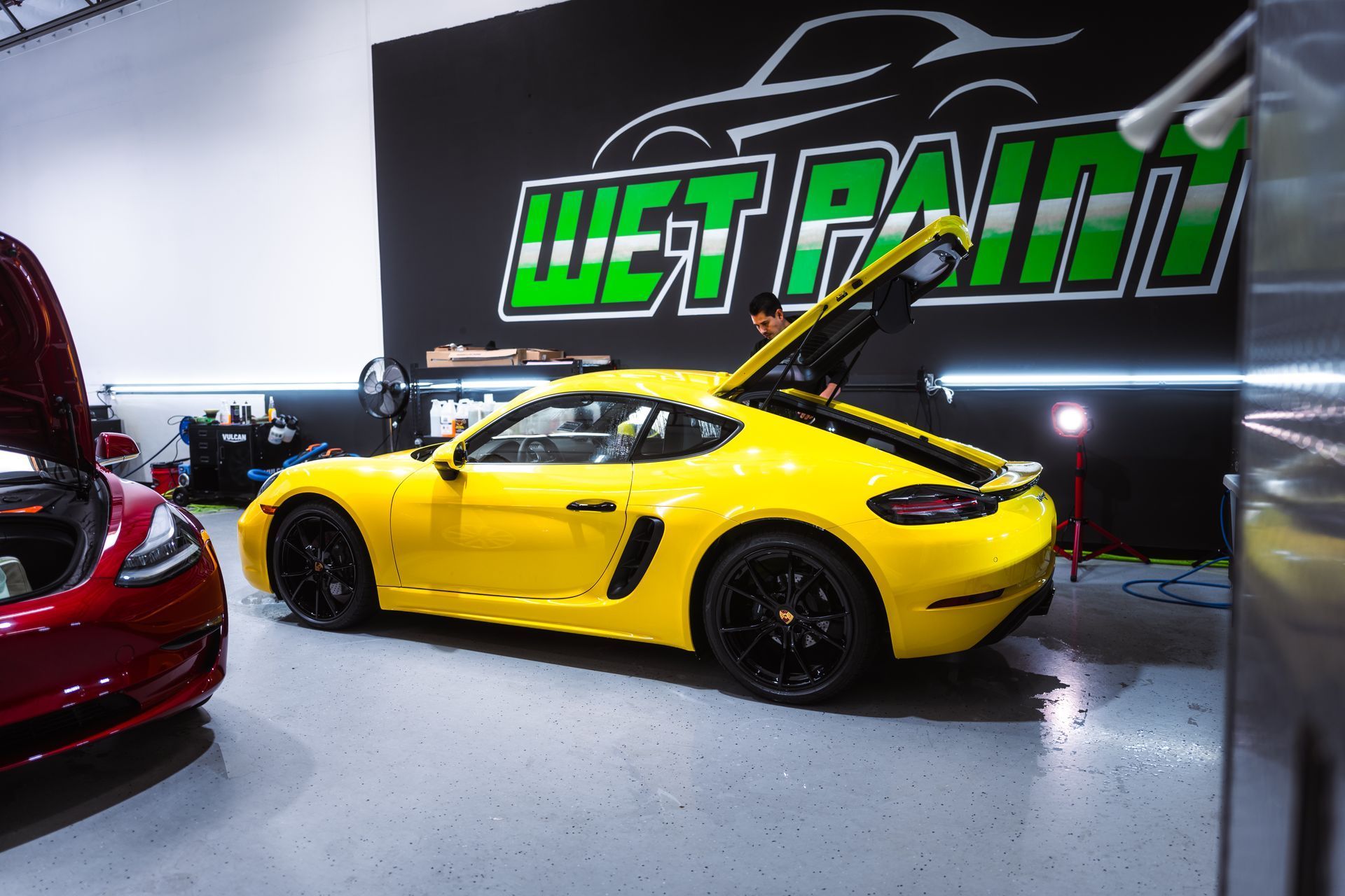 Yellow Porsche with open trunk inside a shop, next to a red car. The shop wall has a 
