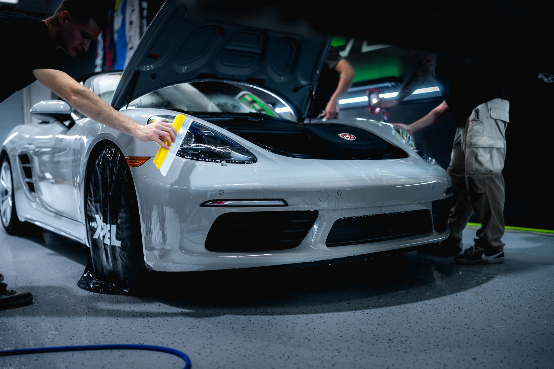 Men applying wrap to a white Porsche's hood in a garage.