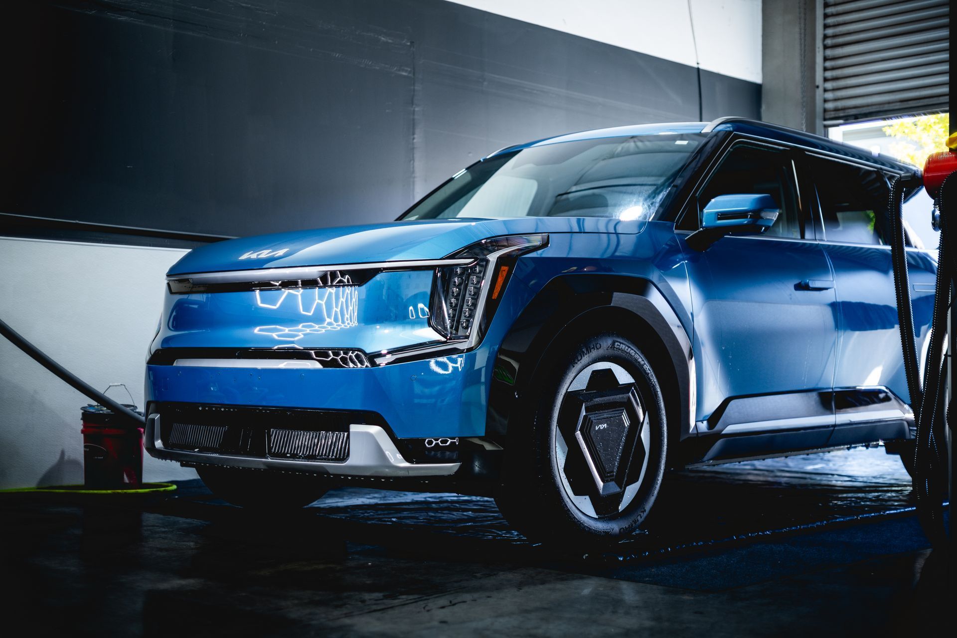 A blue Kia EV9 electric SUV parked inside a garage, highlighting its geometric wheels and modern front headlight design.