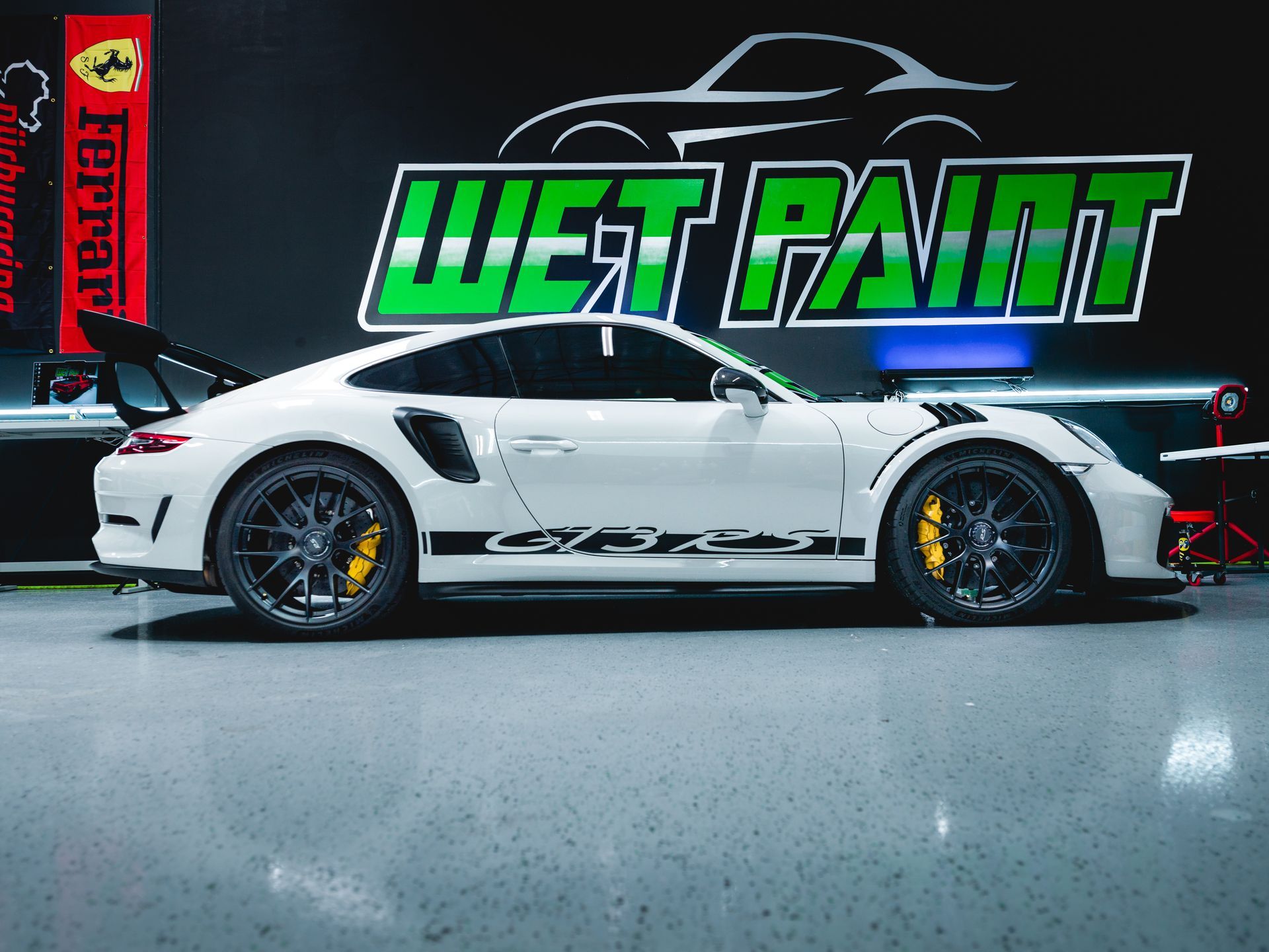 White Porsche 911 with black racing decals in a garage, under a neon “WET PAINT” sign
