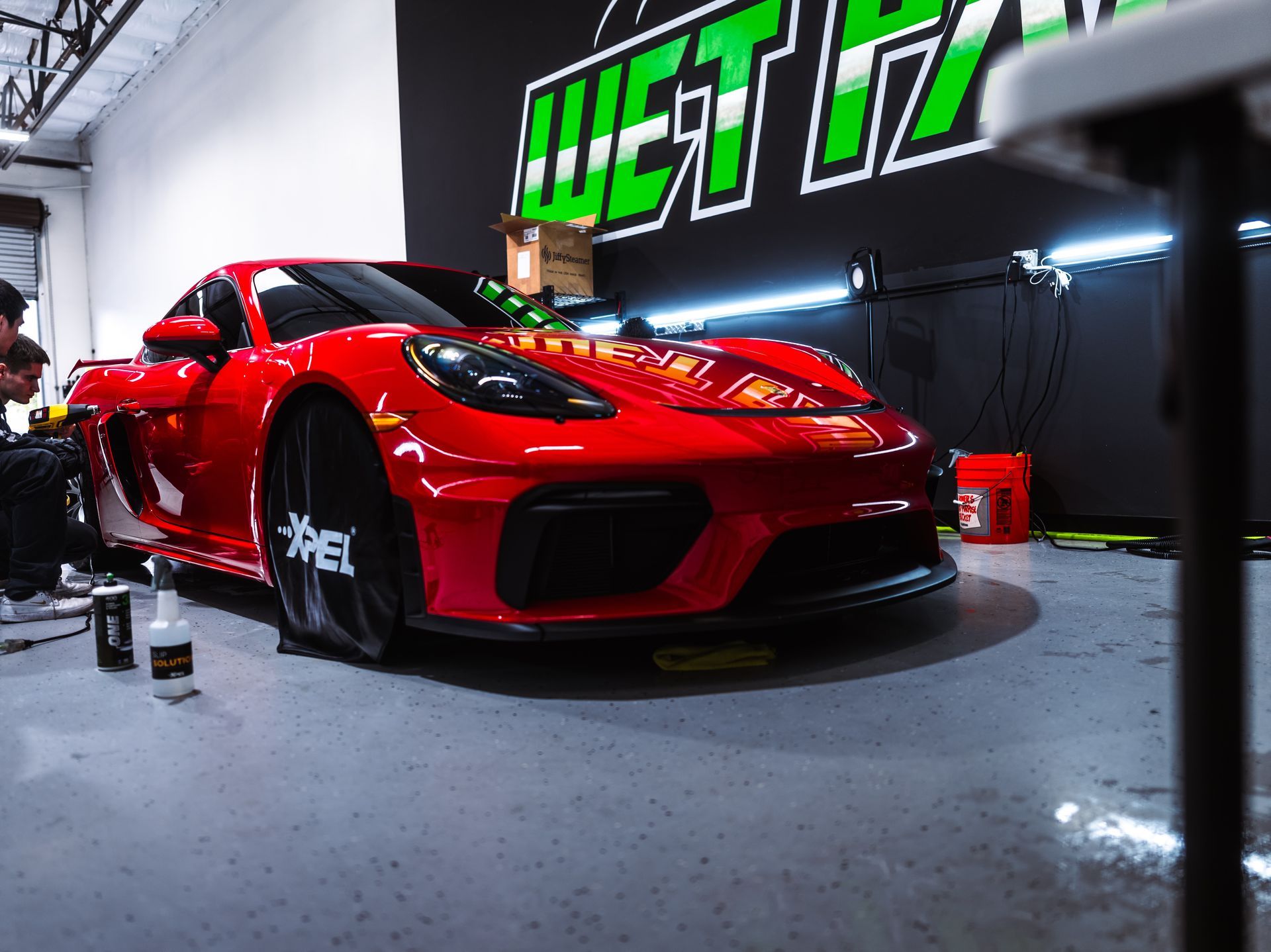 A red sports car is parked in a garage in front of a wet iz sign.