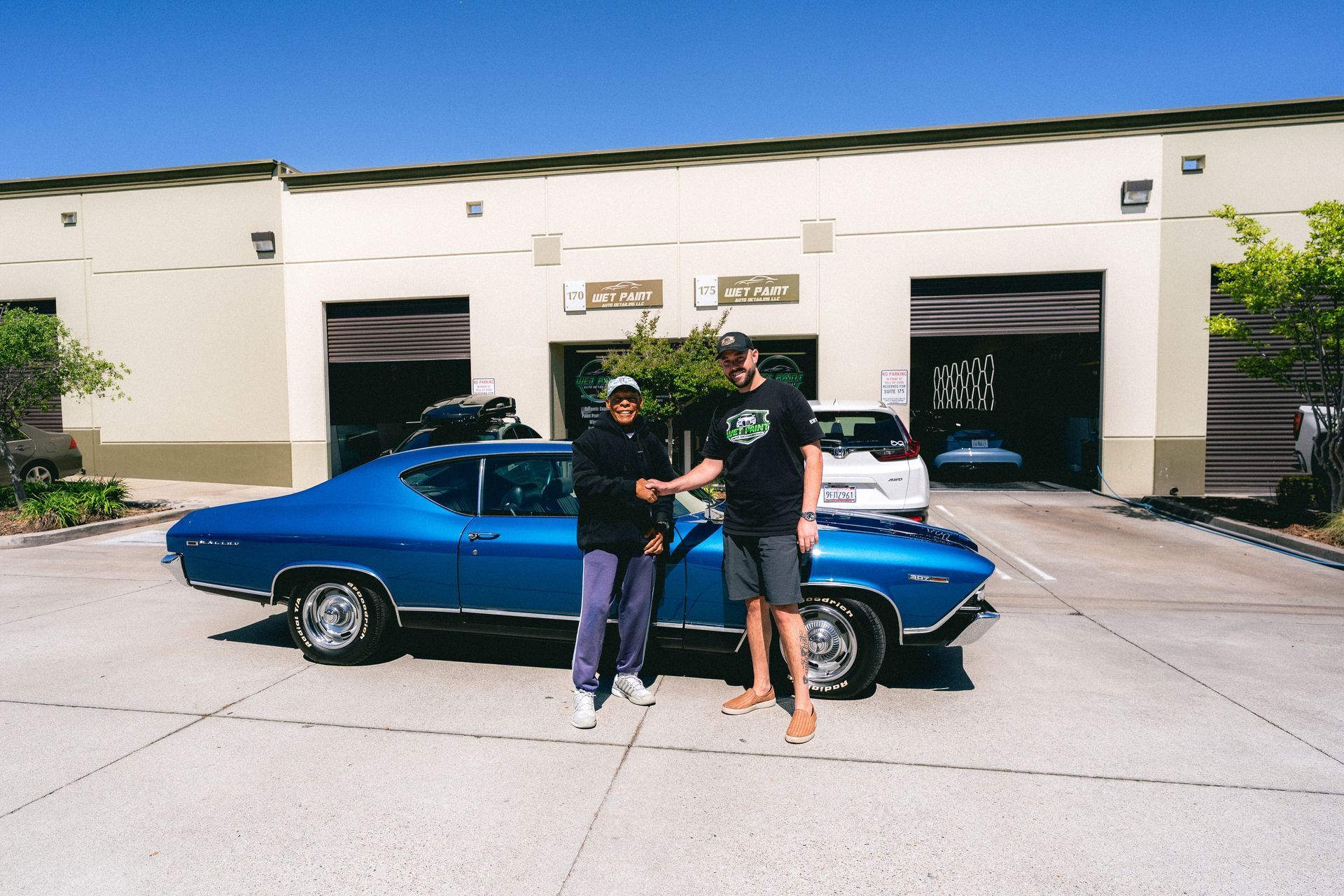 Two people standing beside a blue classic car in front of a building.