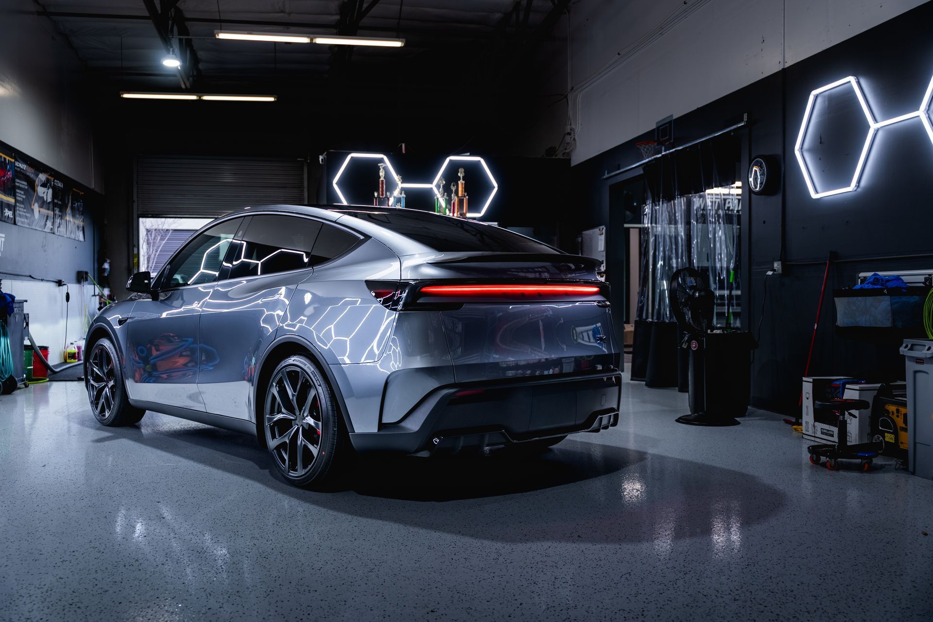 A silver Tesla Model Y parked inside a professional garage with hexagonal wall lights reflecting on its body.