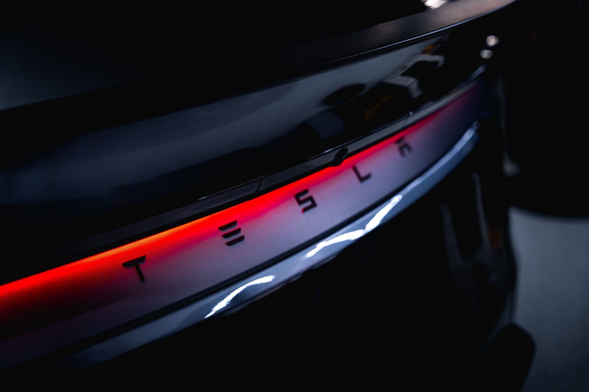 Close-up of a glowing red Tesla rear light bar with the brand name in black lettering against a dark vehicle body.
