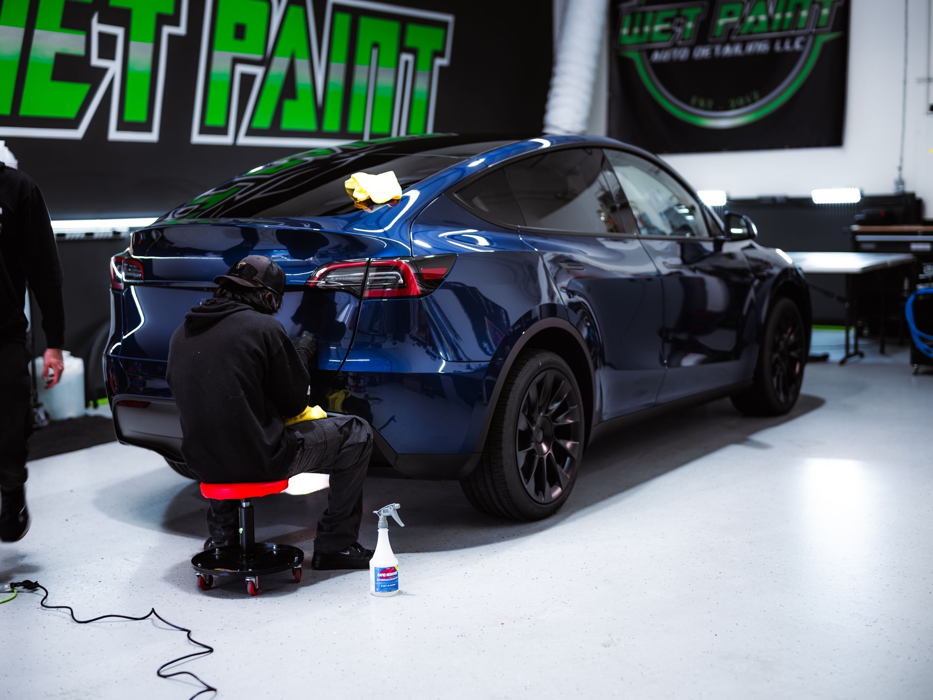 A technician on a rolling stool cleans a dark blue Tesla Model Y in an auto detailing shop.