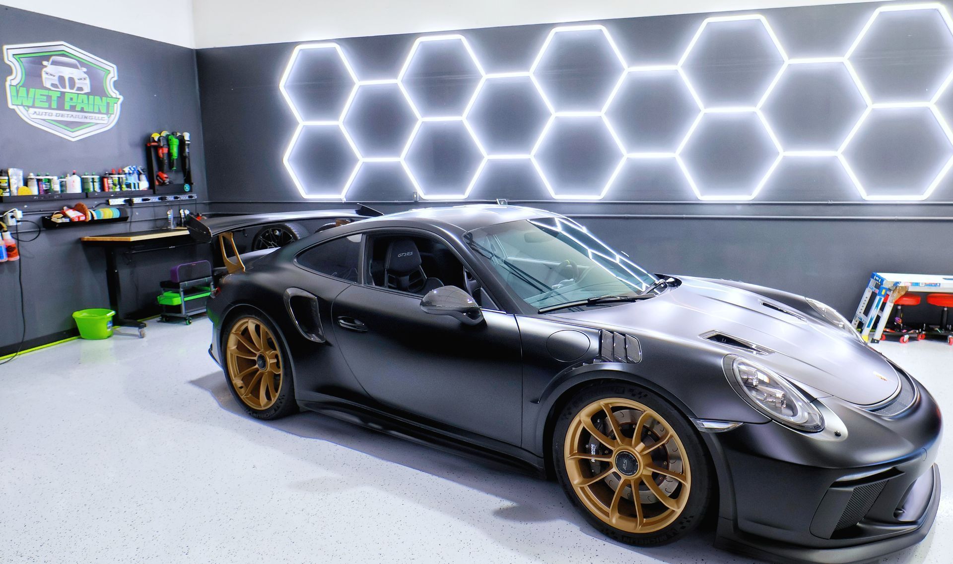 Black Porsche 911 GT3 RS with gold wheels in a detail shop, illuminated by hexagonal lights.