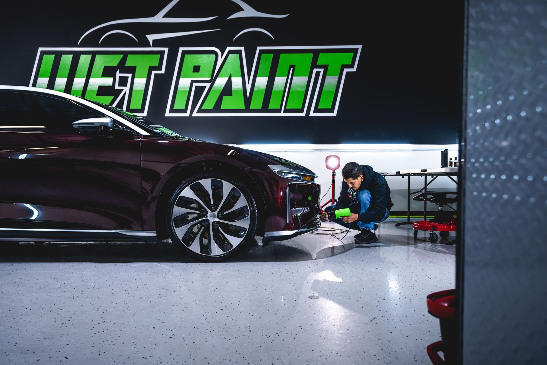 Man working on a burgundy car's front bumper inside a garage with