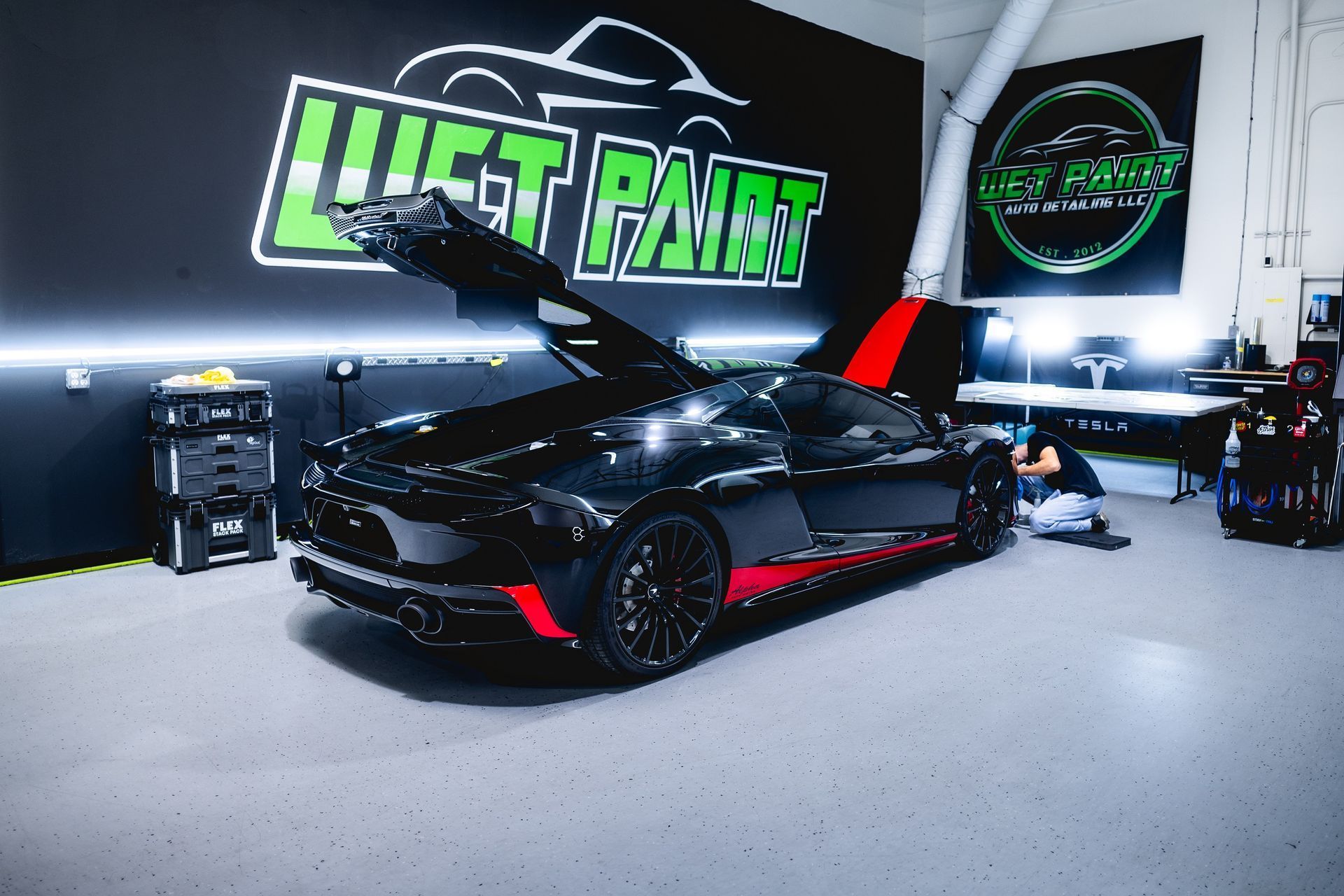 Black sports car with red accents inside a detailing shop, hood open, logo visible.