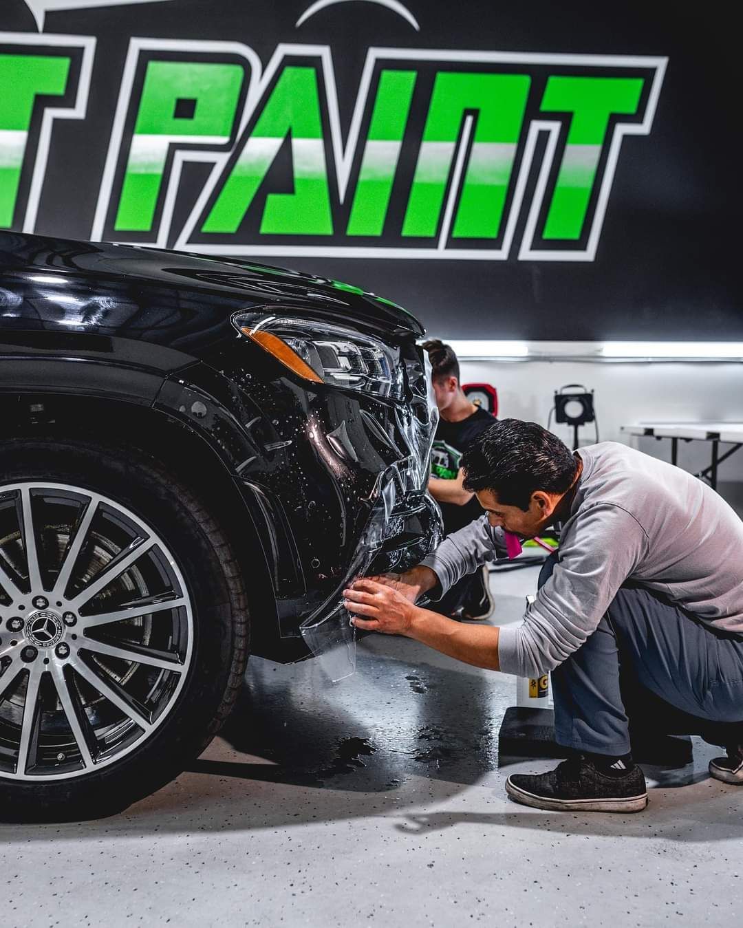 Two men applying a protective film to the front bumper of a black SUV in a garage with bright green and black branding.