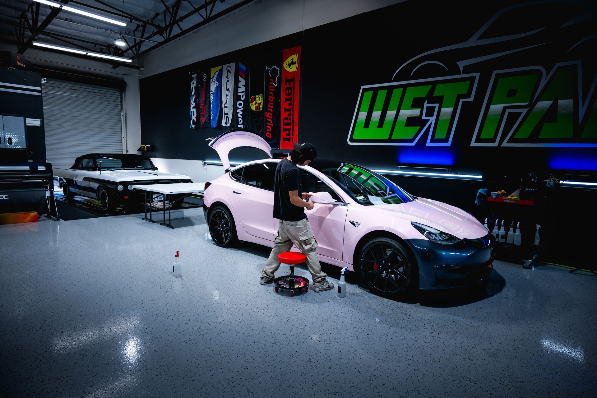 A worker on a stool wraps a light pink Tesla in a bright garage with 