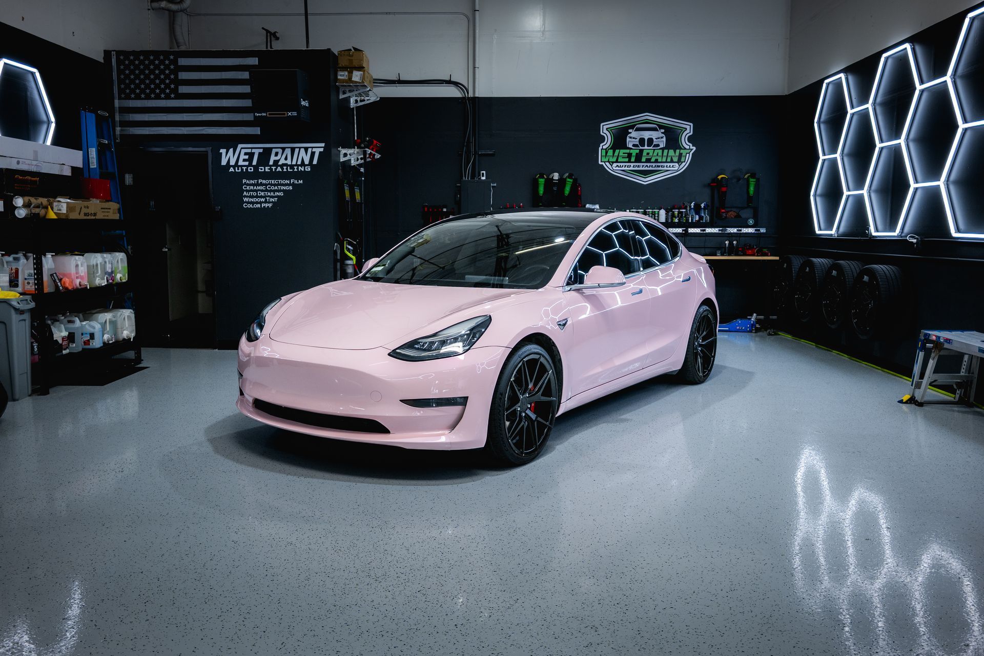 A pale pink Tesla parked in a professional auto detailing garage with hexagon-shaped wall lights and a polished floor.