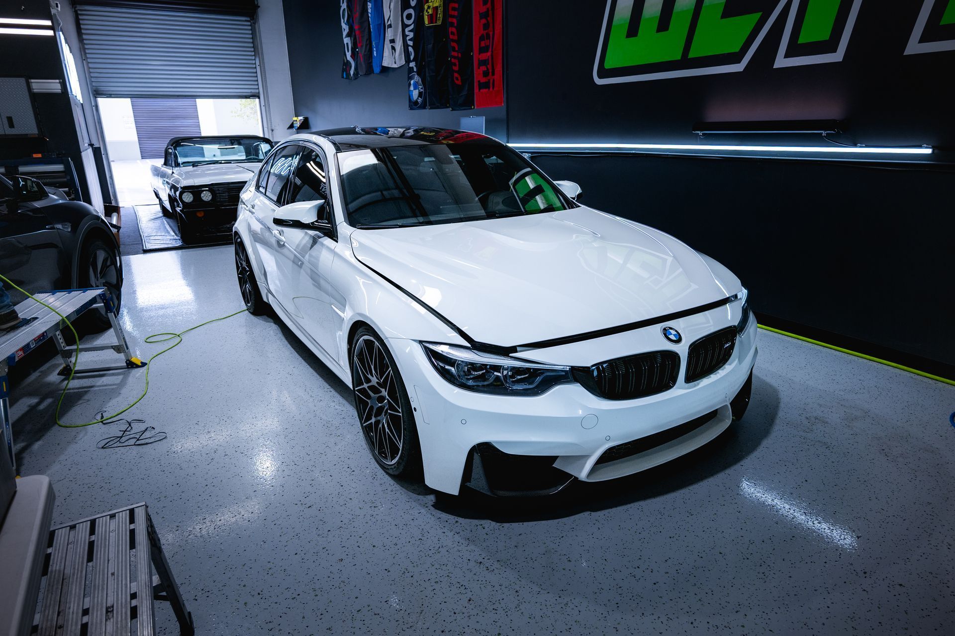 White BMW M3 in a garage, black rims, parked next to other vehicles.
