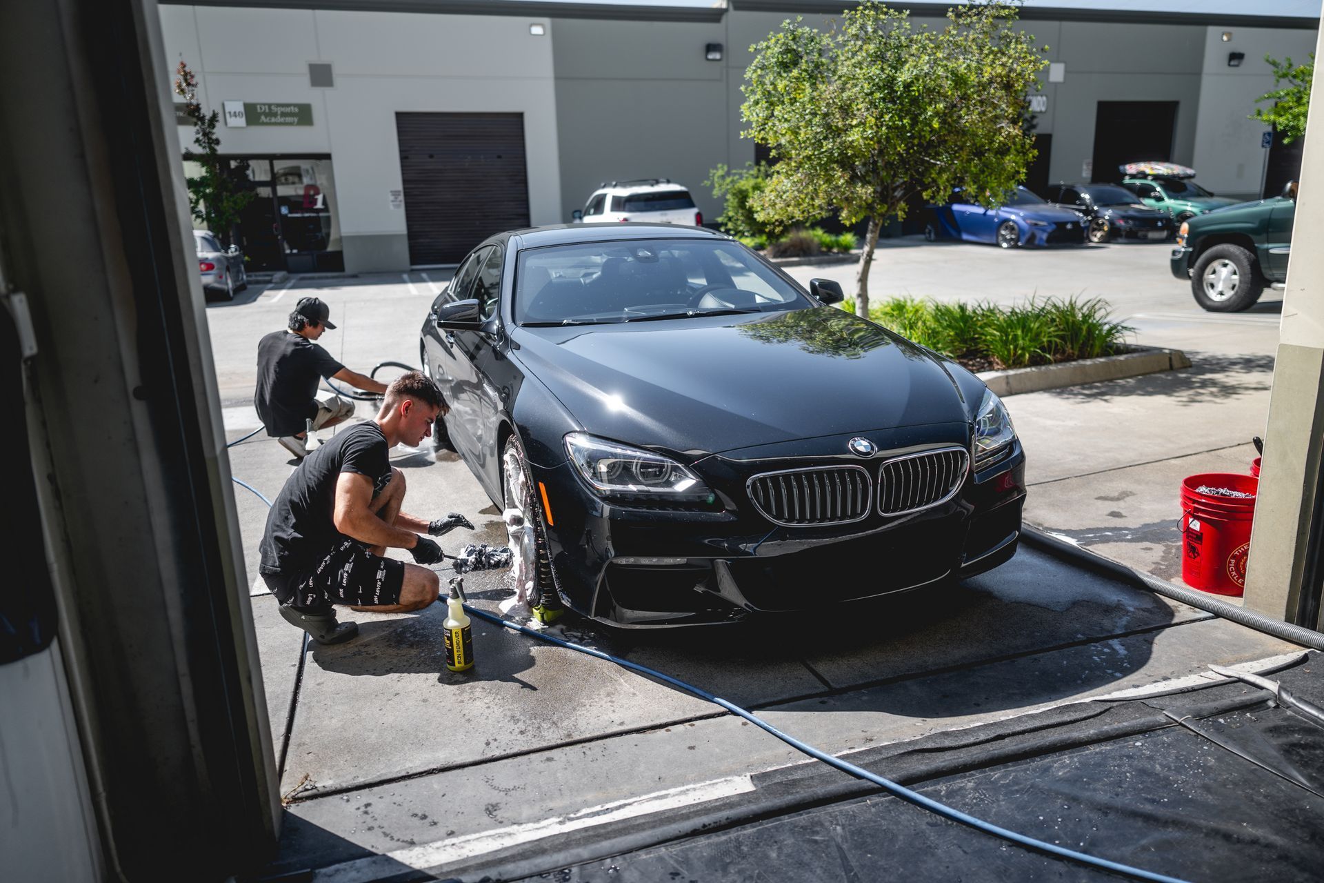 Two people washing a black BMW on a car hauler ramp outside a building.