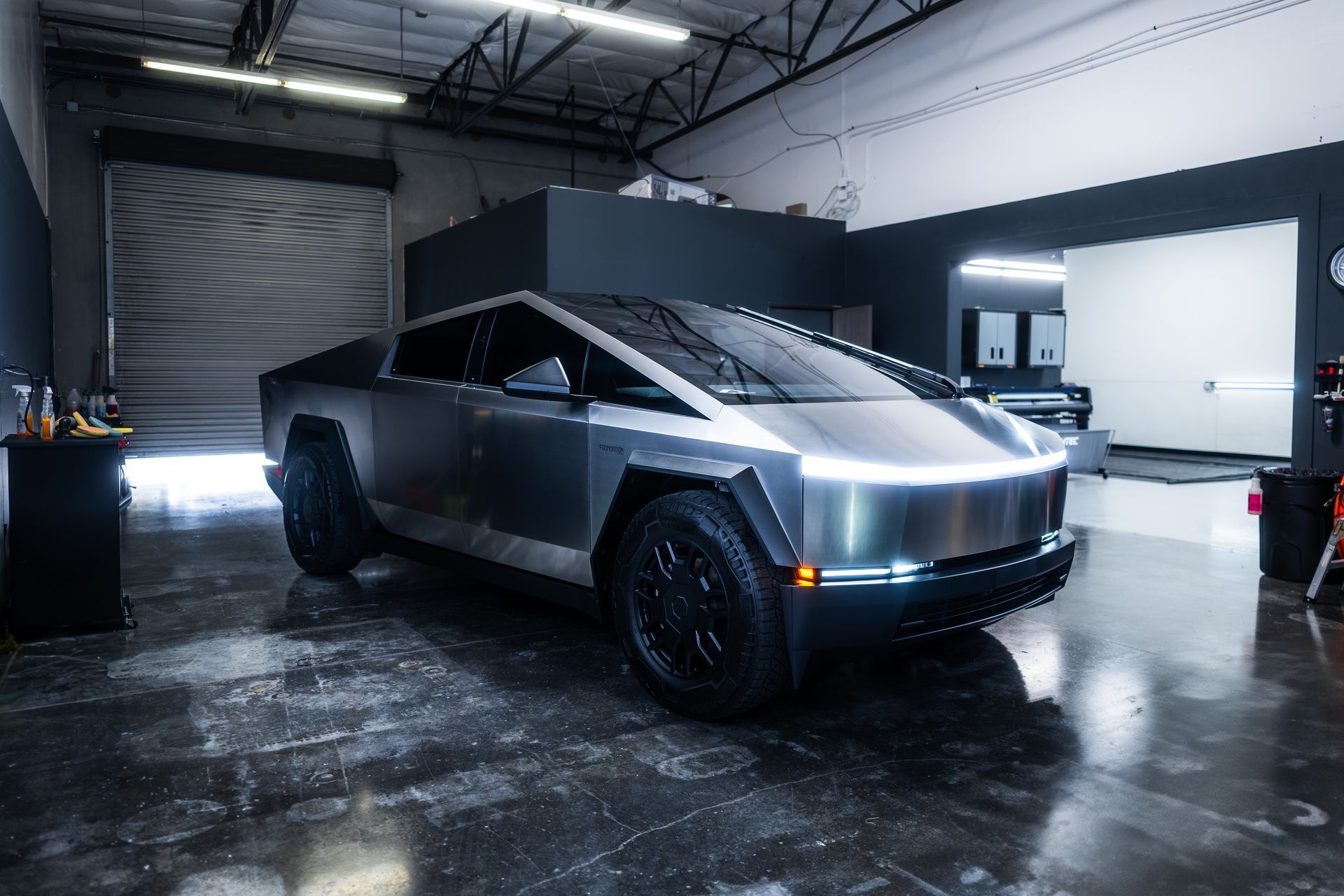 A Tesla Cybertruck parked in a bright, industrial garage with a silver metallic body and black wheels.