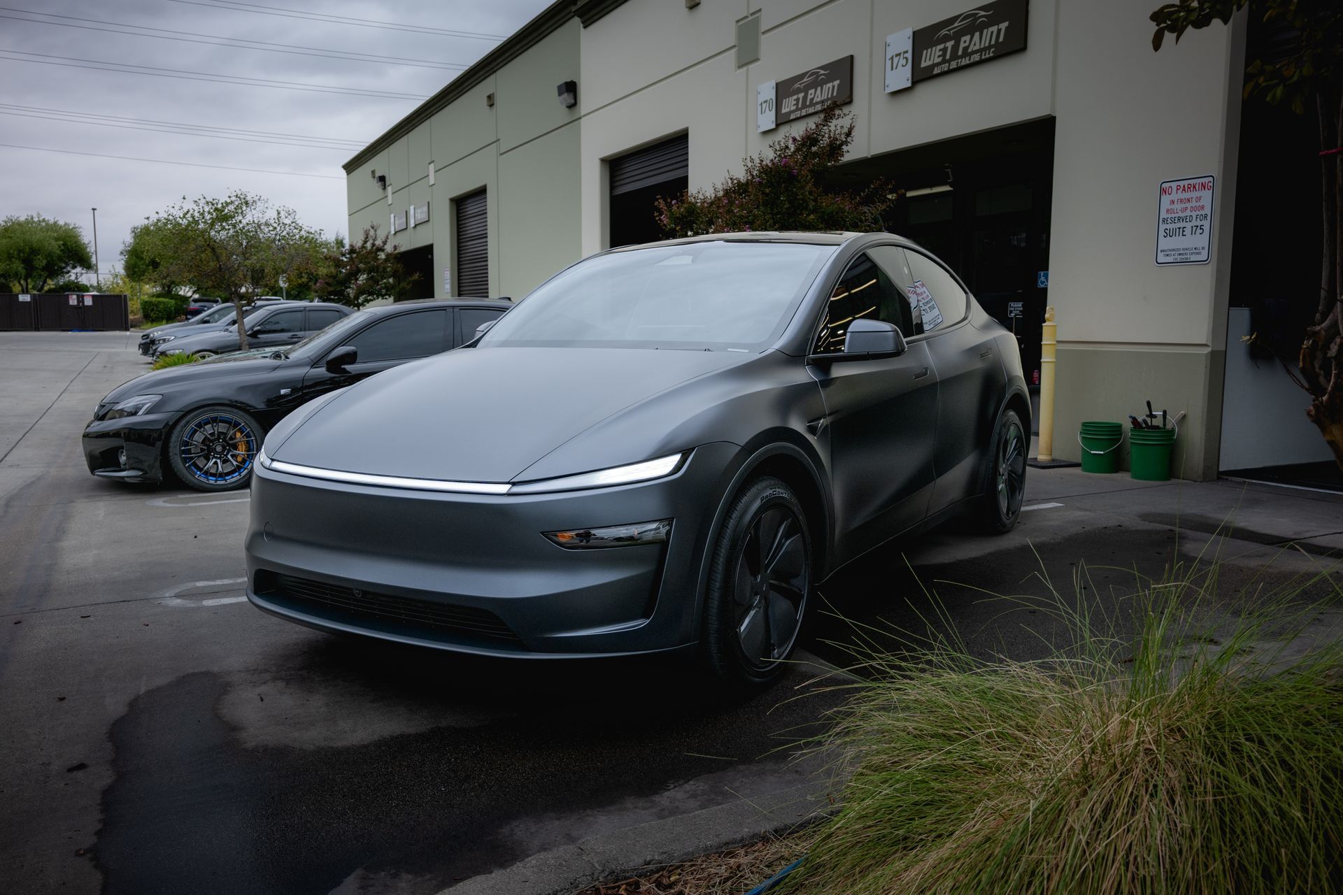 Two dark gray Tesla-style SUVs parked outside a service garage on a wet day