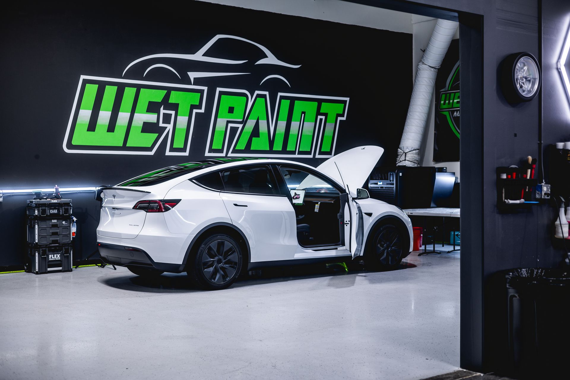 White sports car in a custom garage with an open door and a “WET PAINT” sign on the wall