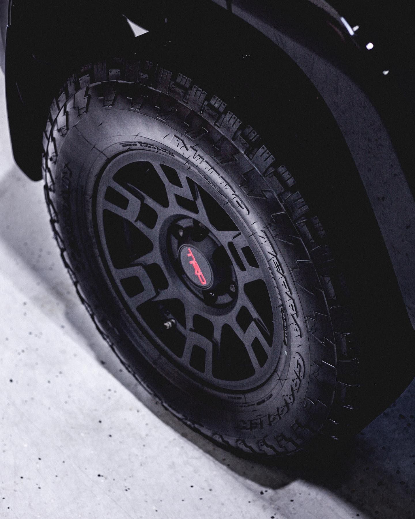 Black off-road vehicle wheel with a black rim and a red TRD logo, on a concrete surface.
