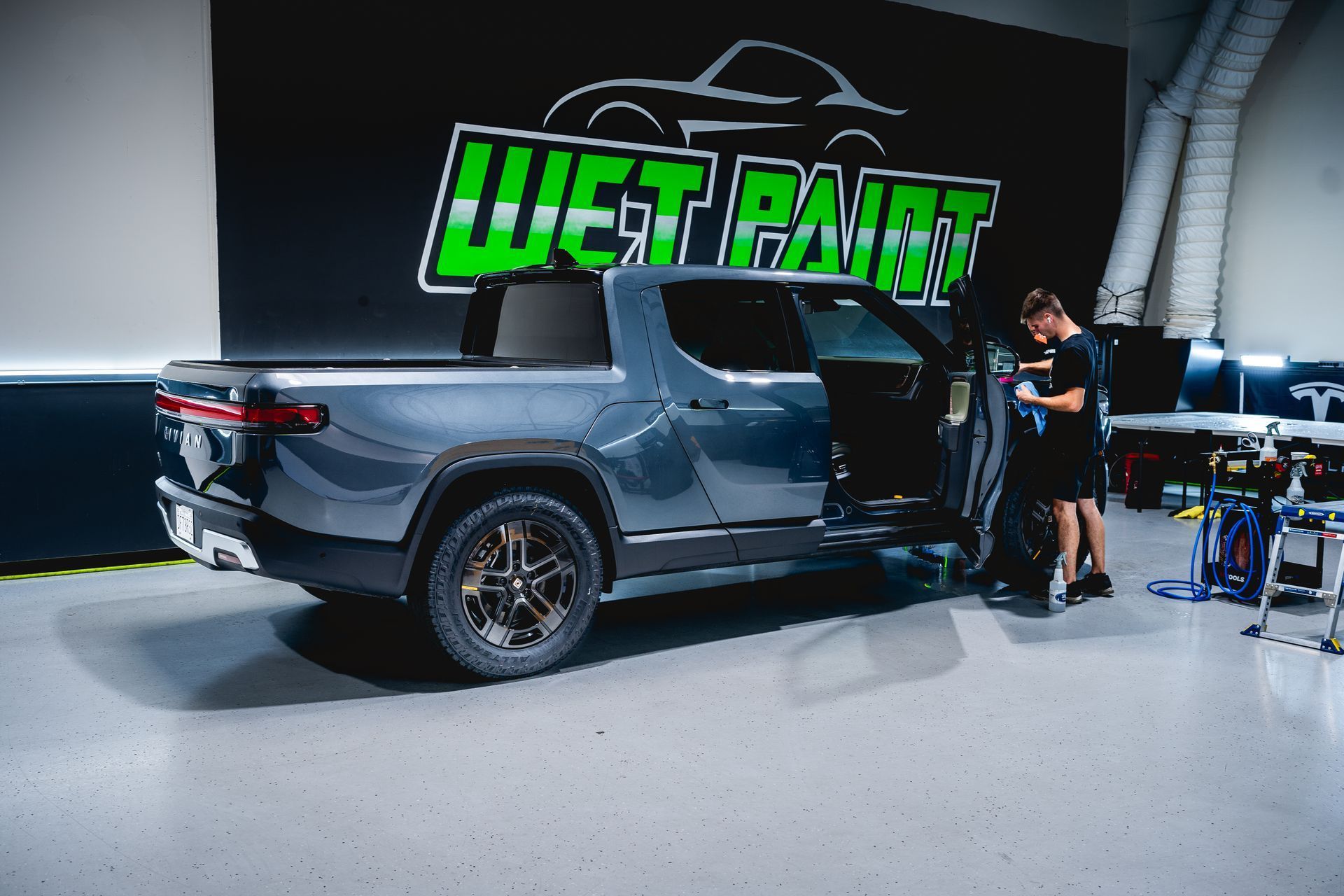 A person works on the door of a dark gray Rivian pickup truck inside a garage with a large 