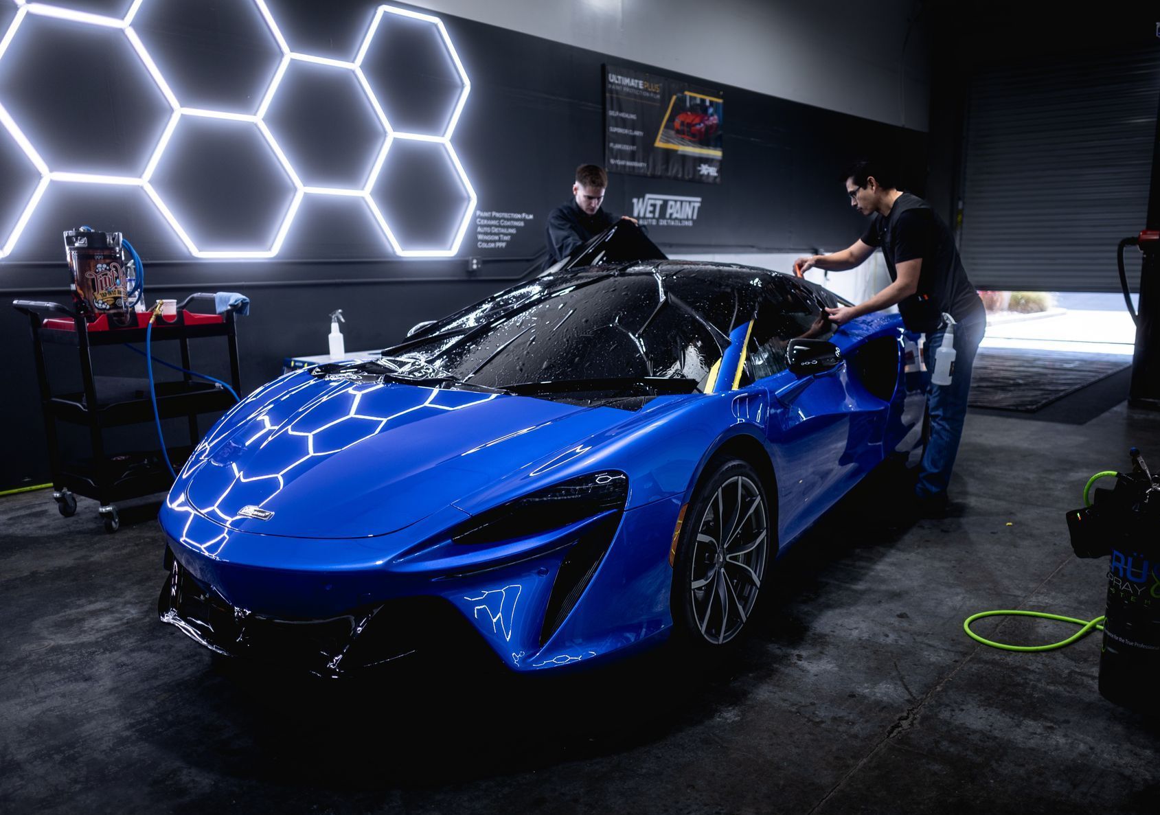 Two men are cleaning a blue sports car in a garage.