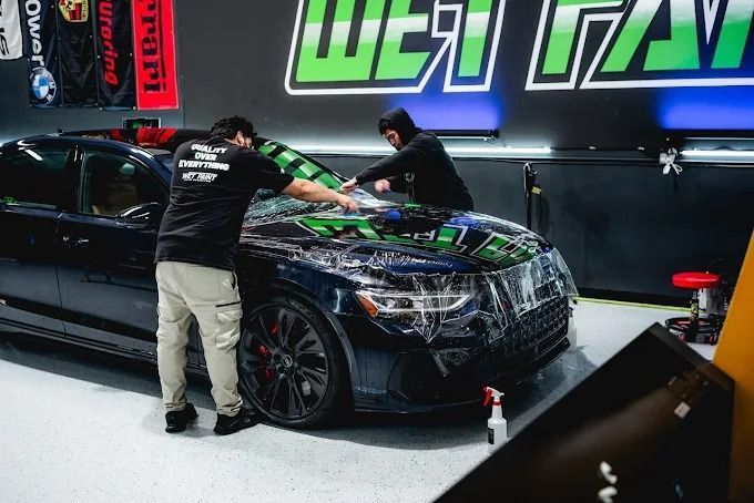 Two Wet Paint technicians applying PPF installation on a dark blue Audi sedan hood in the shop