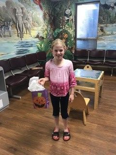 A young girl in a pink and purple shirt and black pants smiles, holding a Halloween bag in a waiting room.