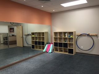 Empty dance studio with large mirror, two cubby shelves, exercise hoops, and colorful rolled mats. 