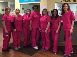 Seven healthcare workers in matching pink scrubs stand behind a check-in desk. 