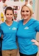 Two women in matching blue shirts smile, standing indoors with colorful decorations on the wall.