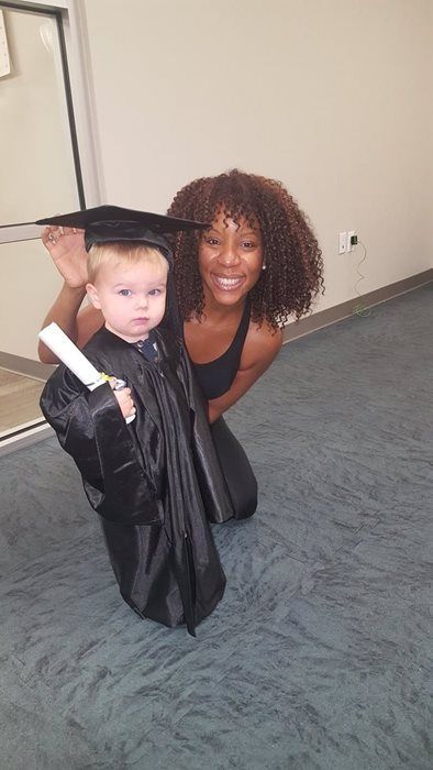 A young child in a graduation gown and cap poses next to a smiling woman. They are indoors on a gray floor.
