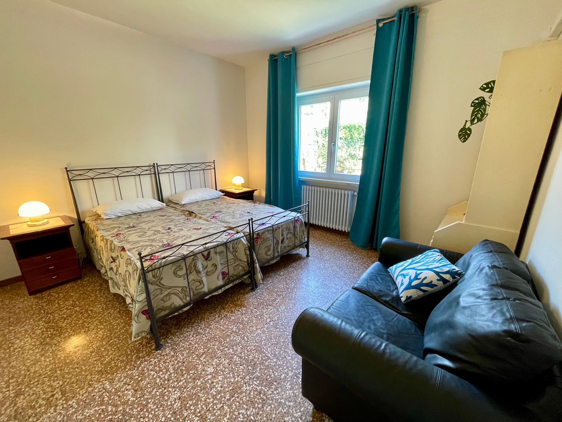 Camera da letto doppia con due letti singoli e due comodini in legno con abatjour vintage, finestra affacciata sul retro e divano nero in pelle