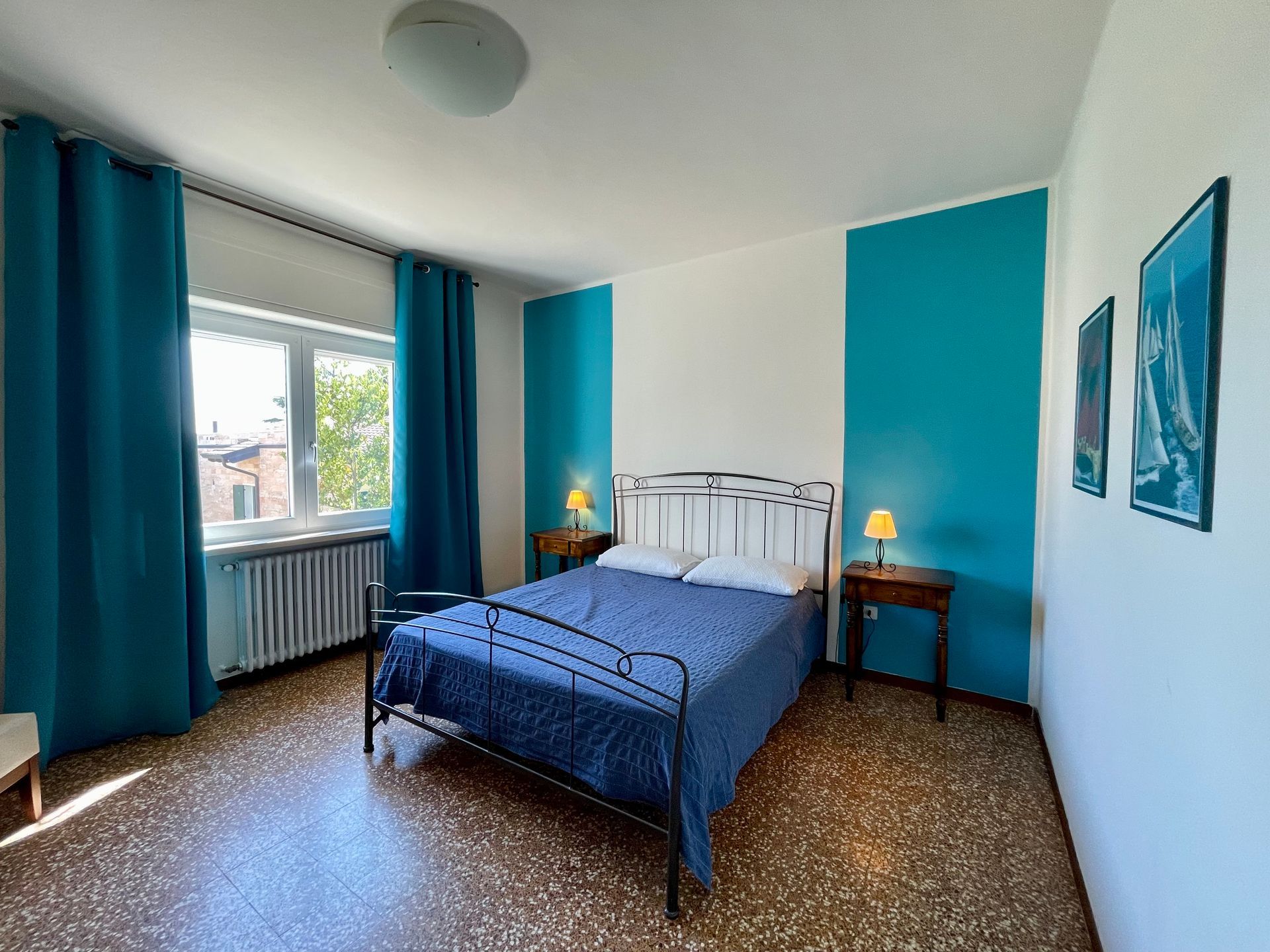 Camera da letto matrimoniale con 2 fasce laterrali8 sul muro dietro il letto in ferro nero con copriletto blu,  due comodini in legno e tende attorno alla finestra vista lago sempre blu